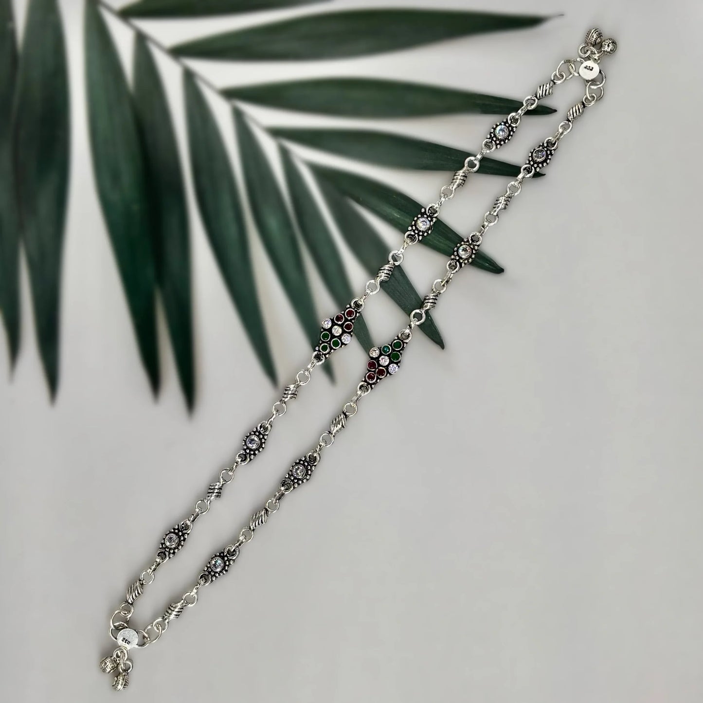 Silver anklet with red, green, and clear gemstones on white background with green palm leaf