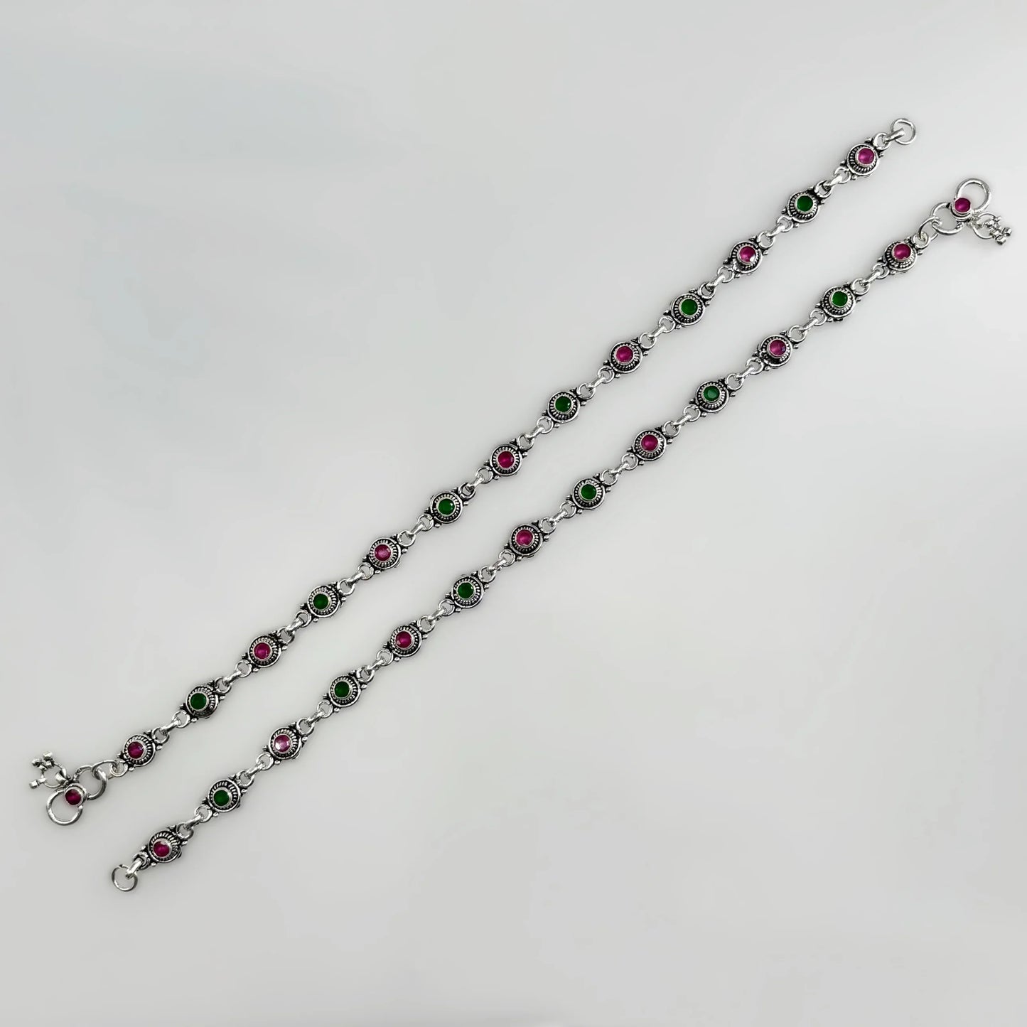 Two silver anklets with alternating green and pink stone settings on a white background