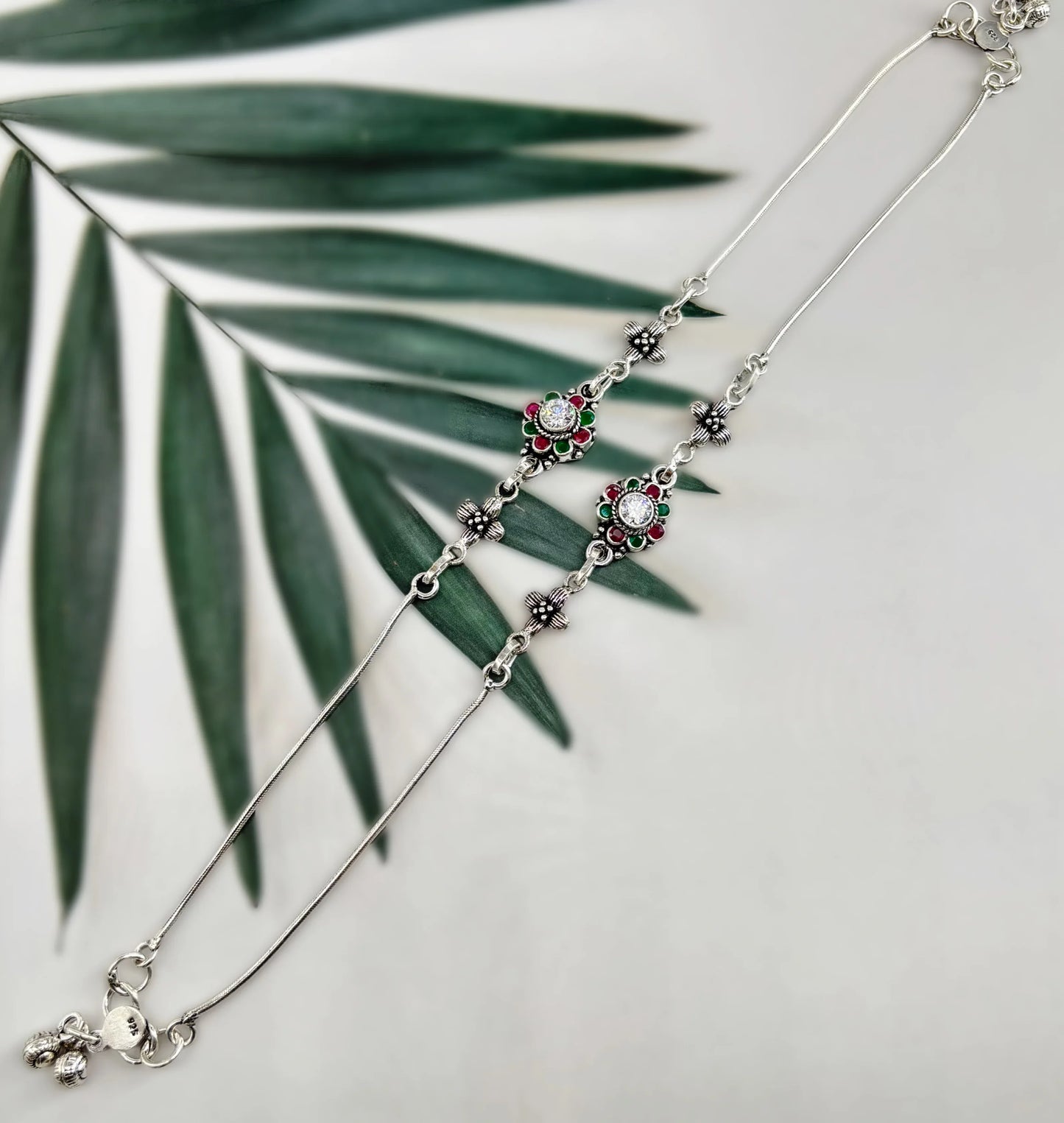 Silver anklet with floral and gem designs in red and green, displayed on green leaves