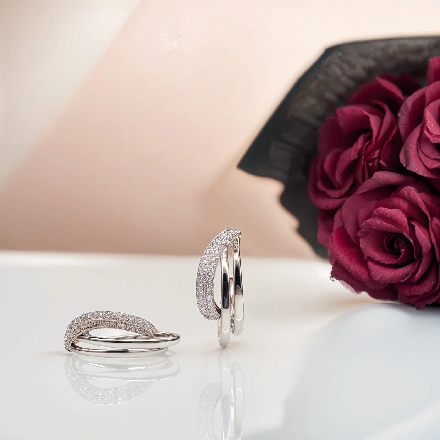 Elegant silver diamond-studded rings on reflective surface with dark red roses background