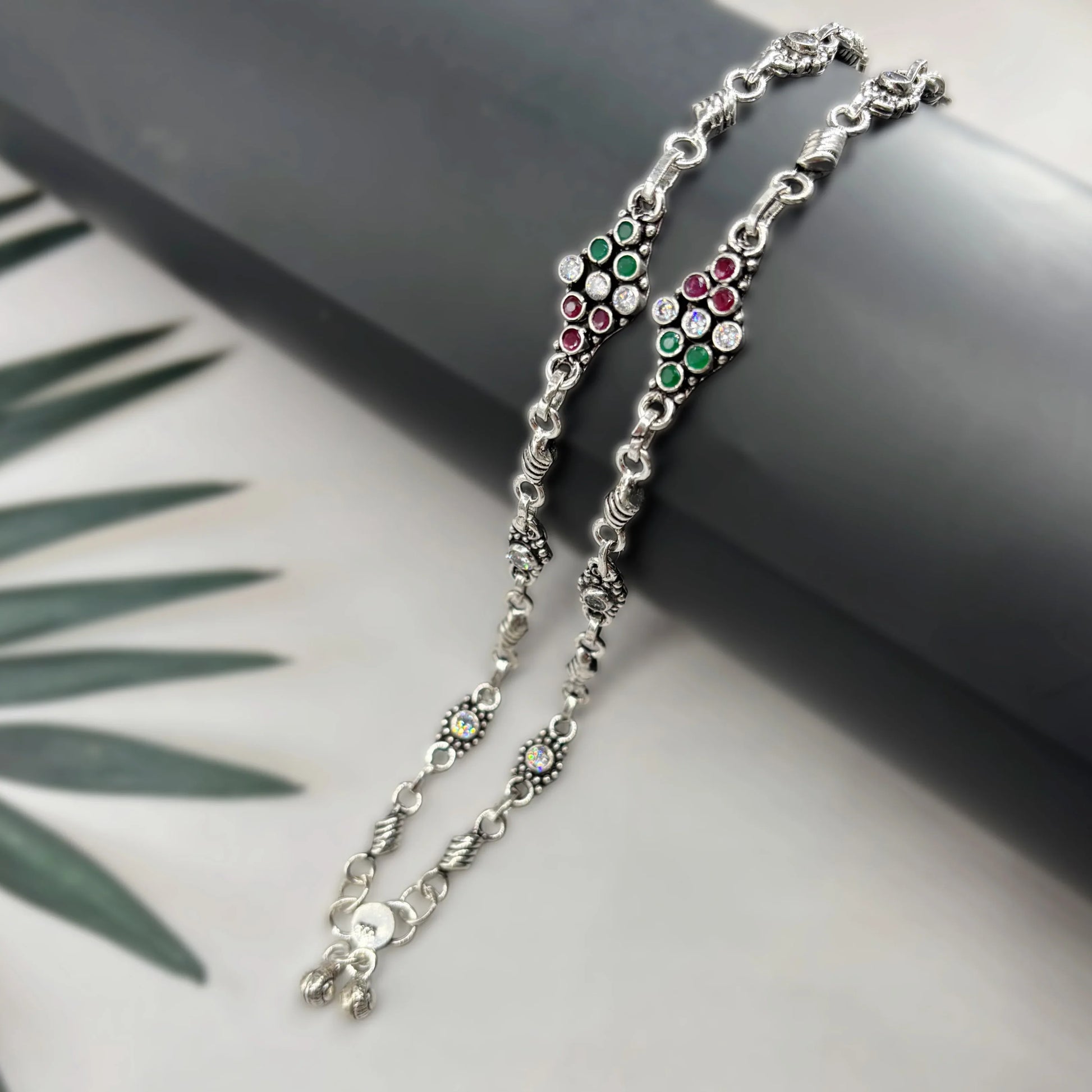 Silver anklets with red and green stones and intricate detailing on black and white background
