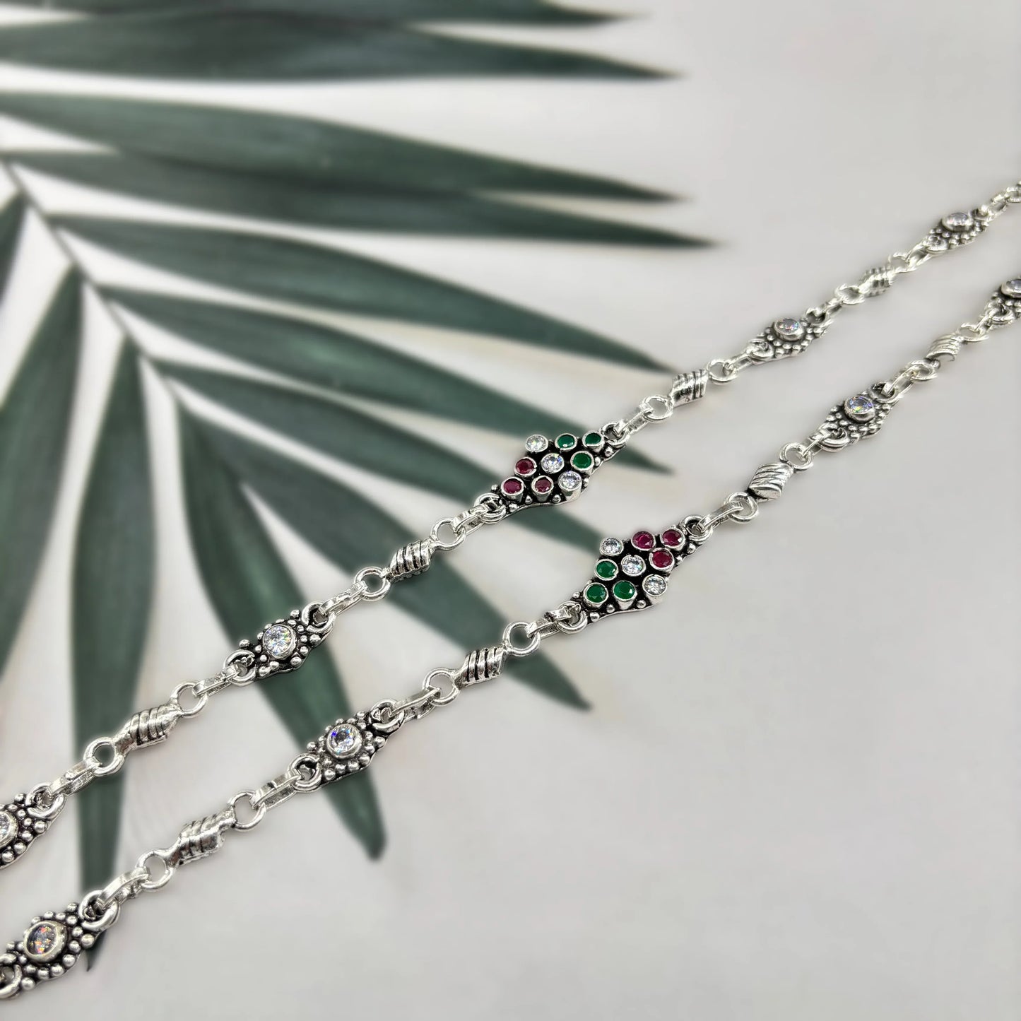 Silver bracelet with red, green, and white gemstones on gray background with green palm leaves