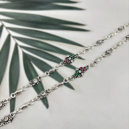Silver bracelet with red, green, and white gemstones on gray background with green palm leaves