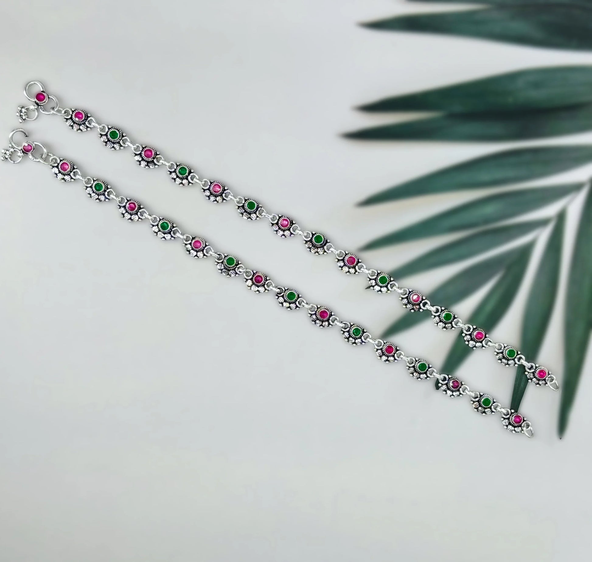 Silver anklets with alternating pink and green round stones on white surface with green leaf