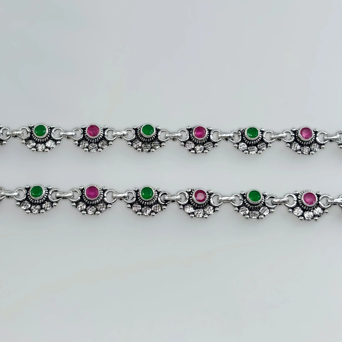Silver anklets with alternating green and pink stones in decorative settings on white background