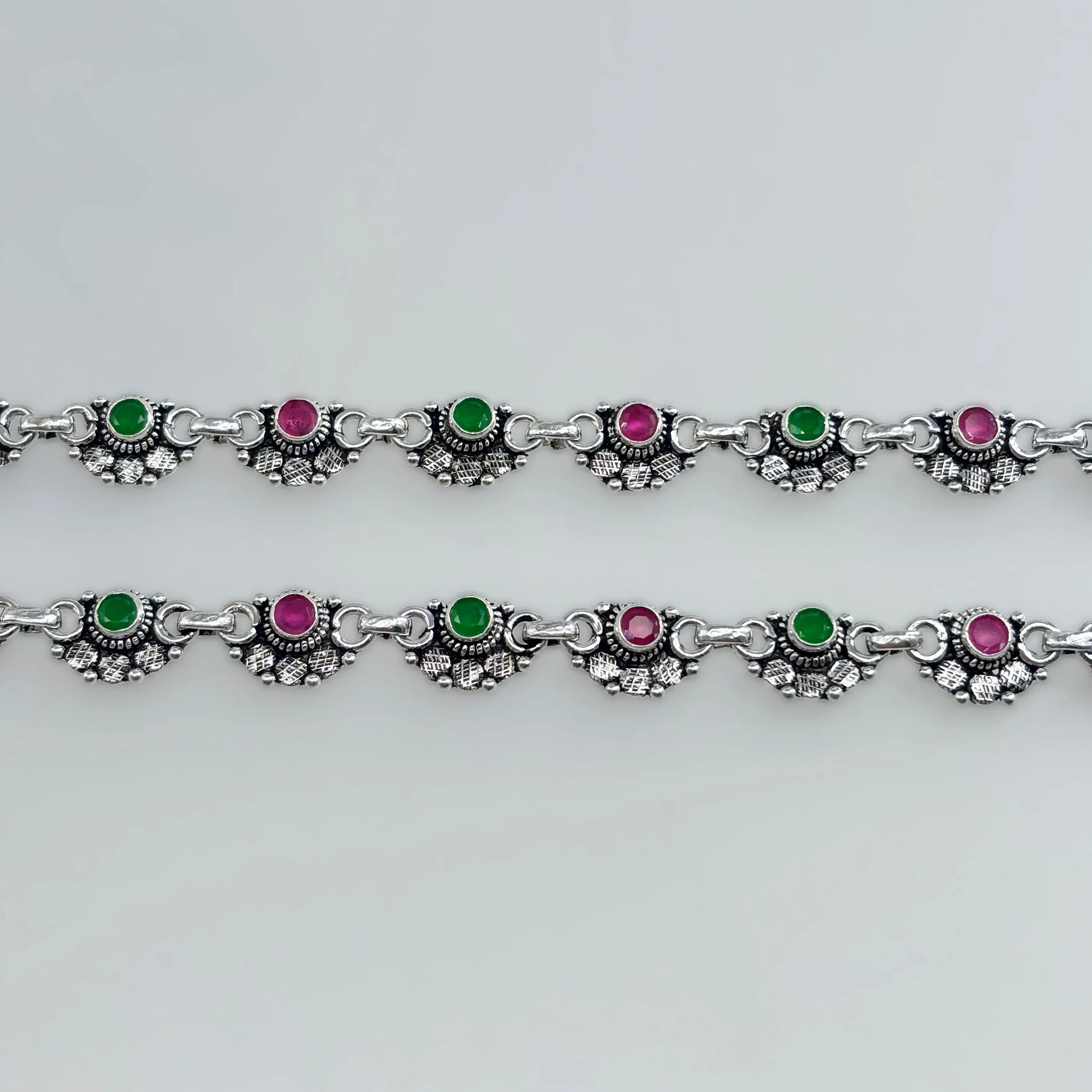 Silver anklets with alternating green and pink stones in decorative settings on white background