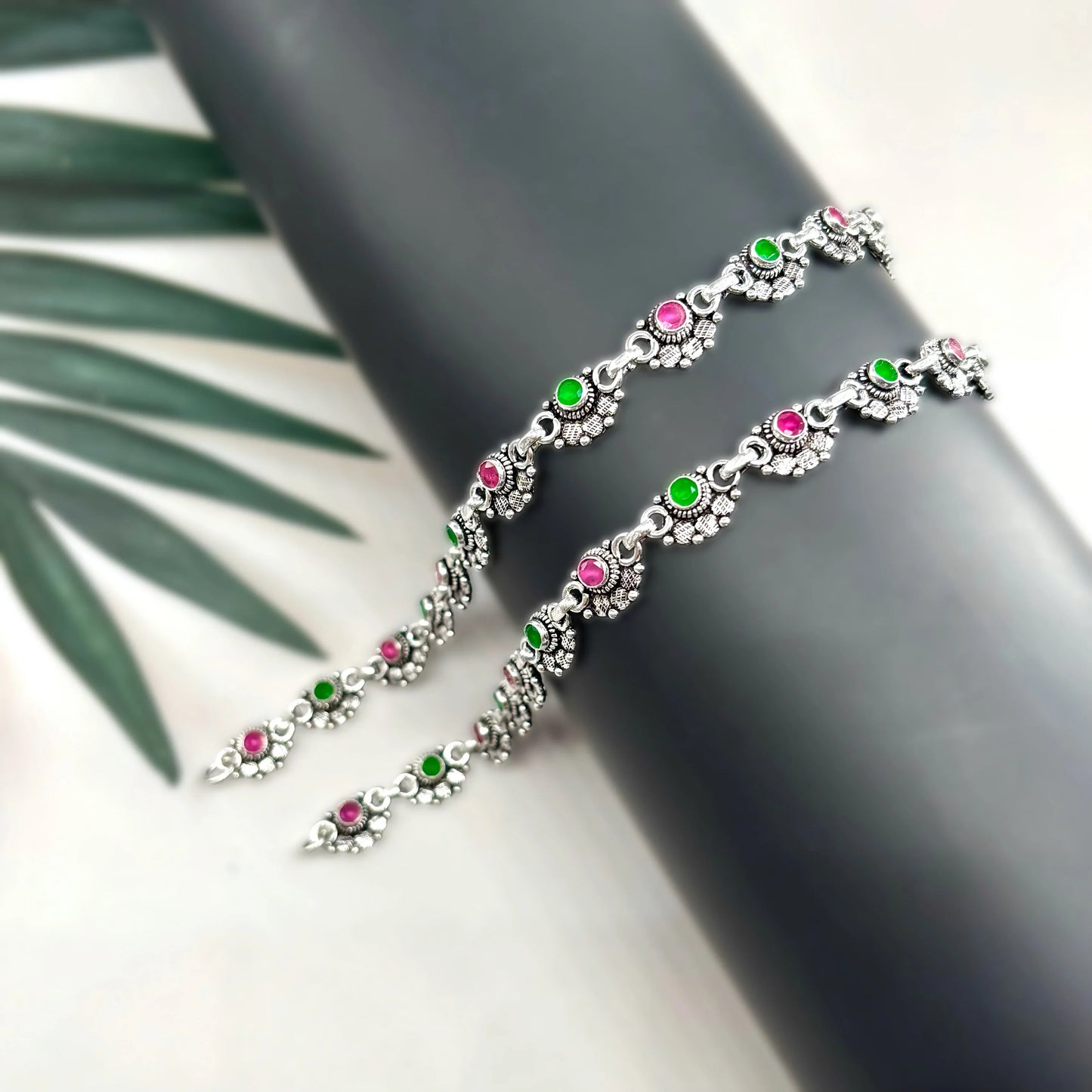 Silver anklets with pink and green stones on black cylindrical stand, decorative jewelry