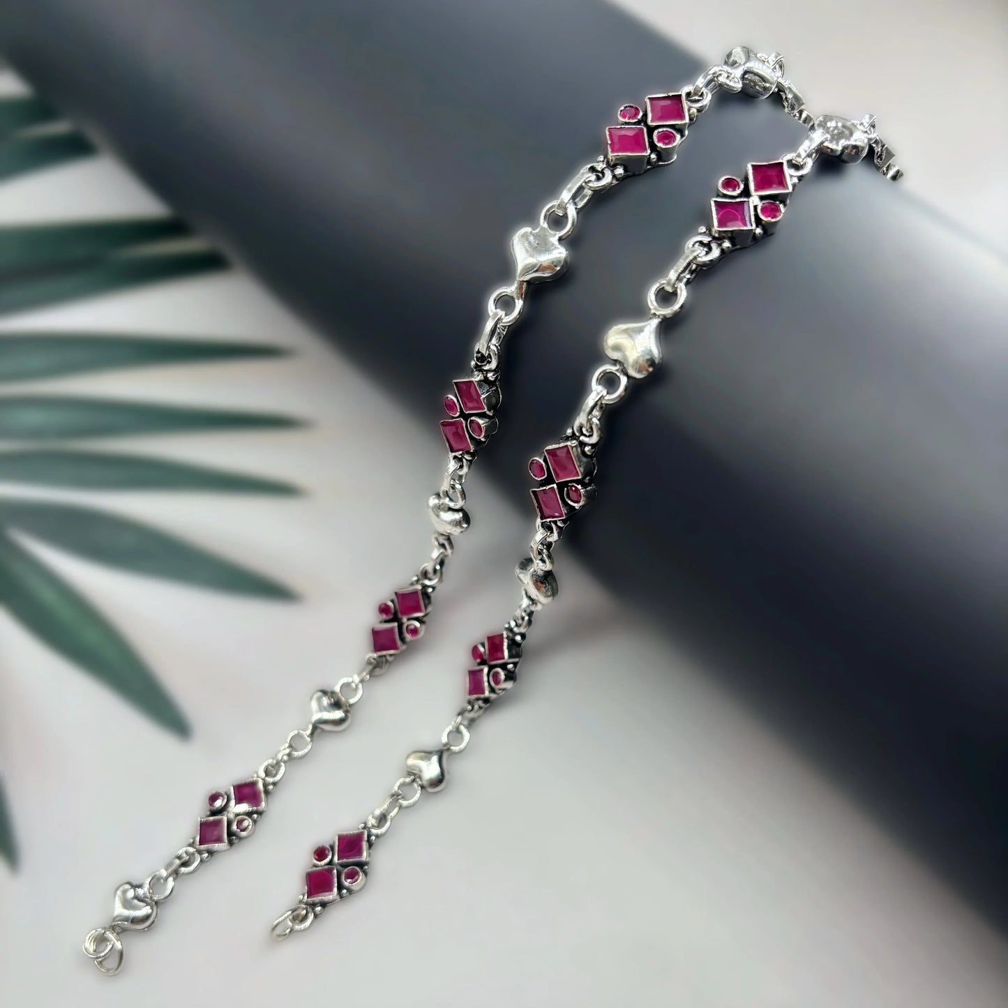 Silver anklets with pink geometric and heart-shaped charms on black cylindrical display