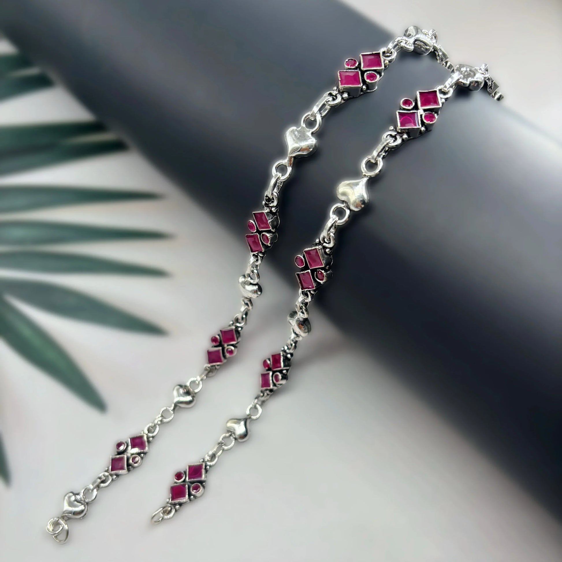 Silver anklets with pink geometric and heart-shaped charms on black cylindrical display