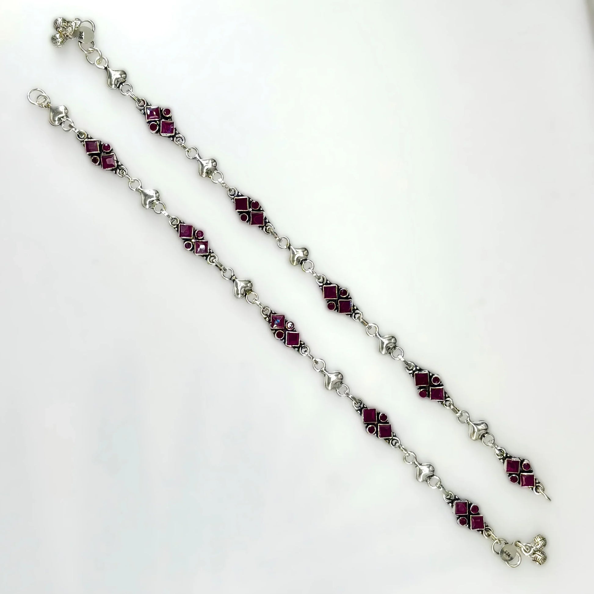 Pair of silver anklets with red square gemstones on white background