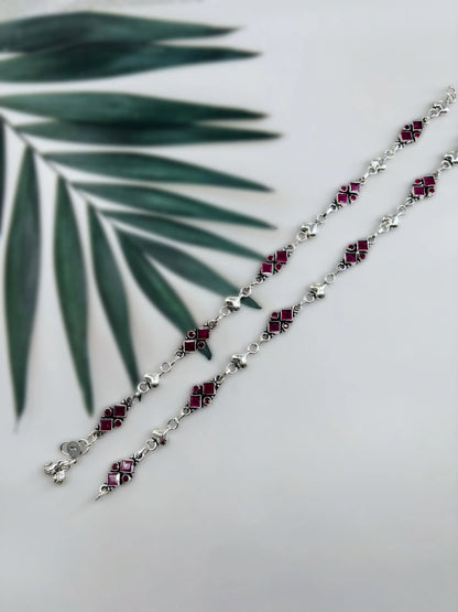Silver anklets with red square gemstones and heart-shaped links on white background with green palm leaf