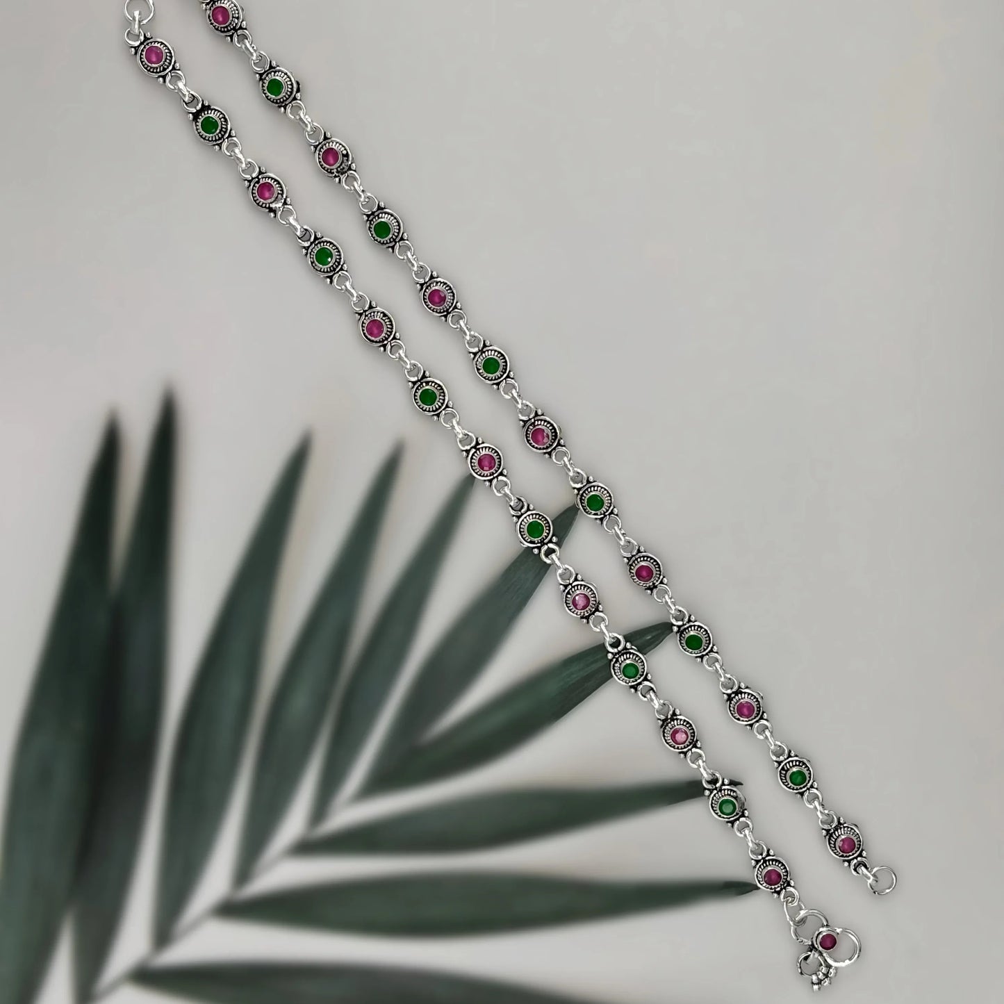 Silver bracelet with alternating green and pink circular stones on gray background with palm leaf