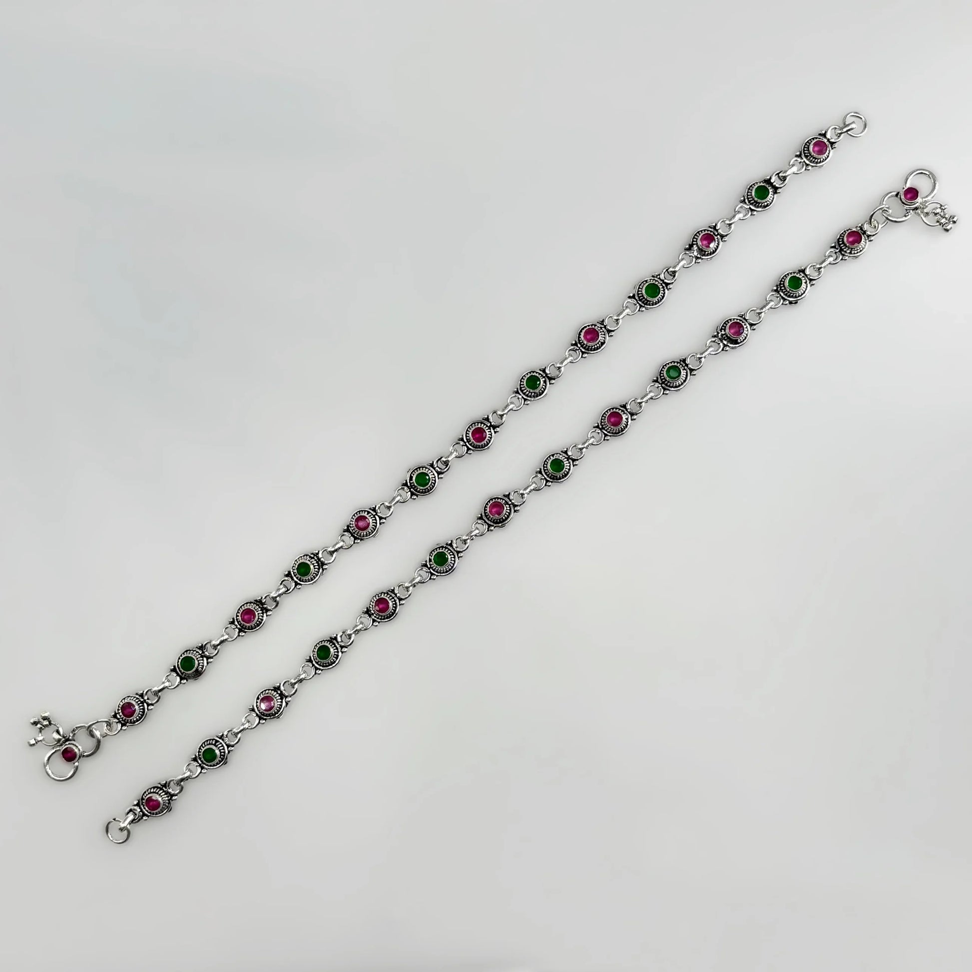 Two silver anklets with alternating green and pink stone settings on a white background