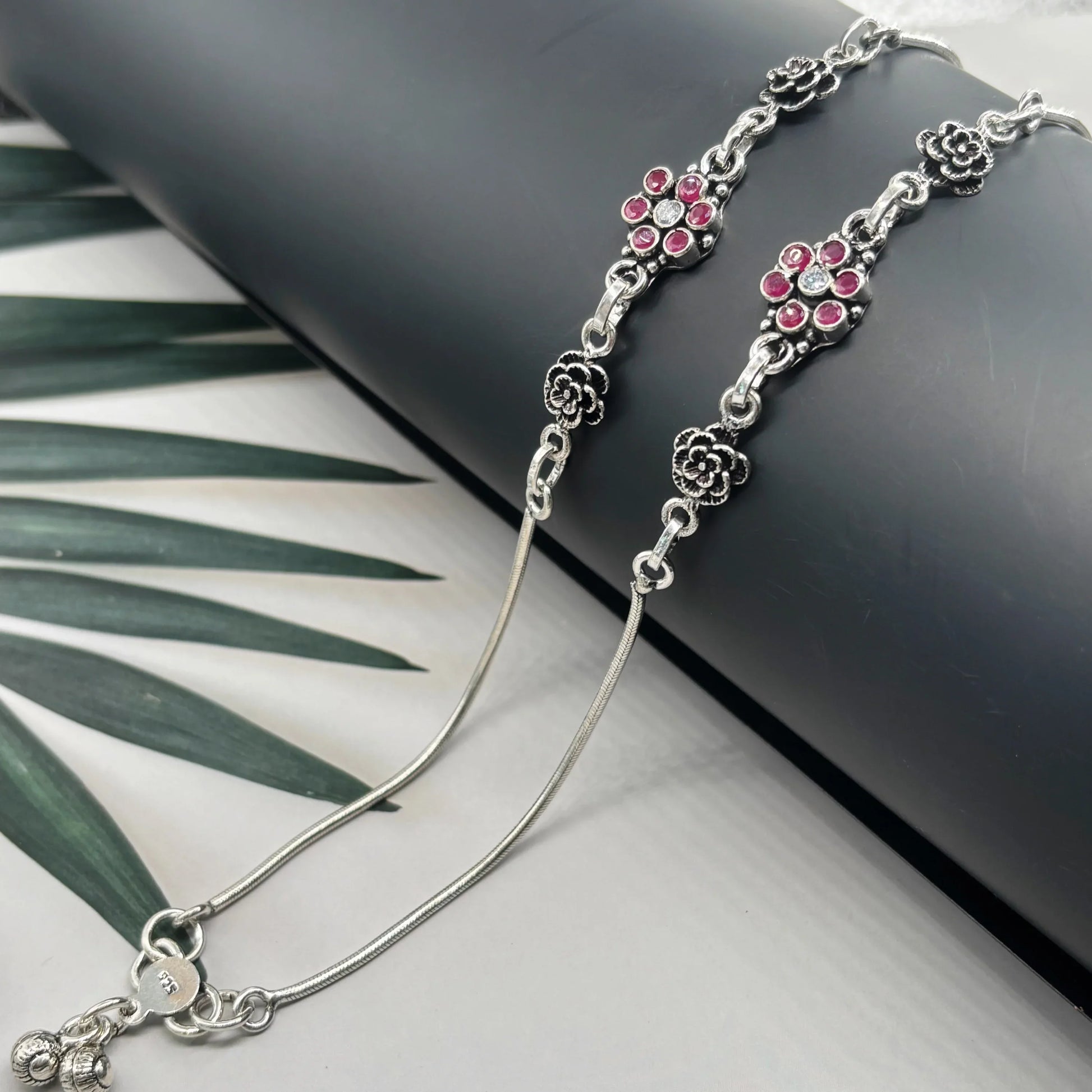 Sterling silver anklets with floral designs and red gemstone accents on a black display