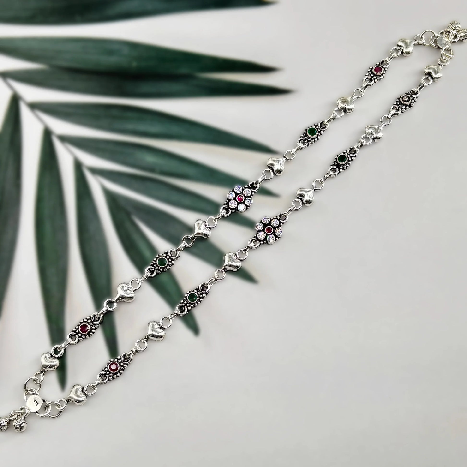 Silver anklet with heart charms, red and green stones, floral design, on white background with green palm leaf