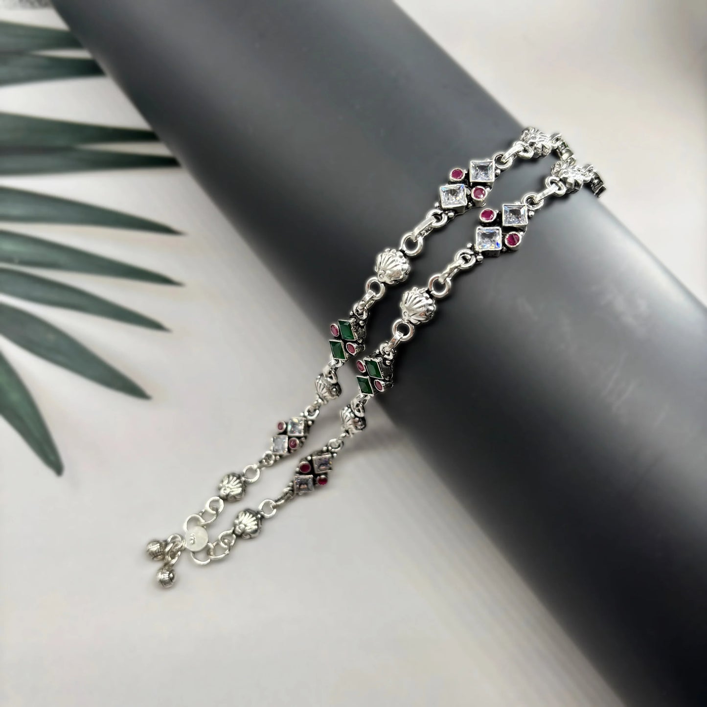 Silver anklets with green and red stones displayed on black cylindrical stand with palm leaves