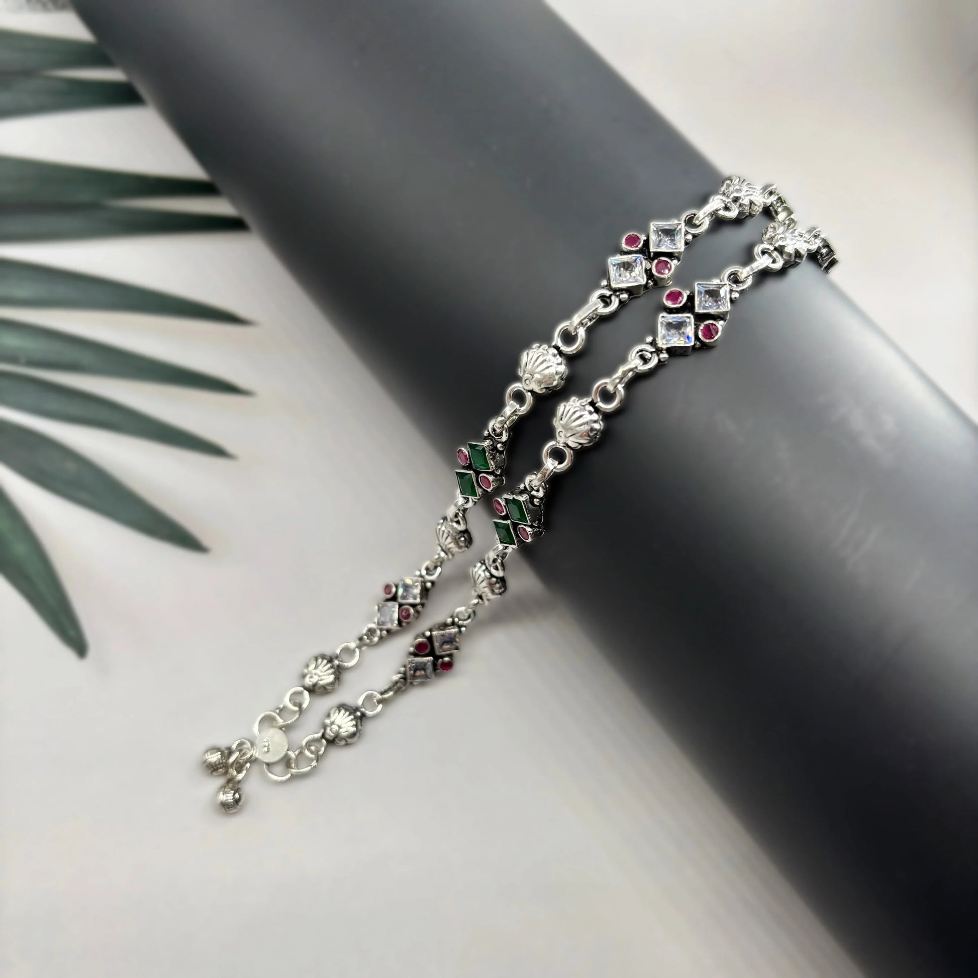 Silver anklets with green and red stones displayed on black cylindrical stand with palm leaves