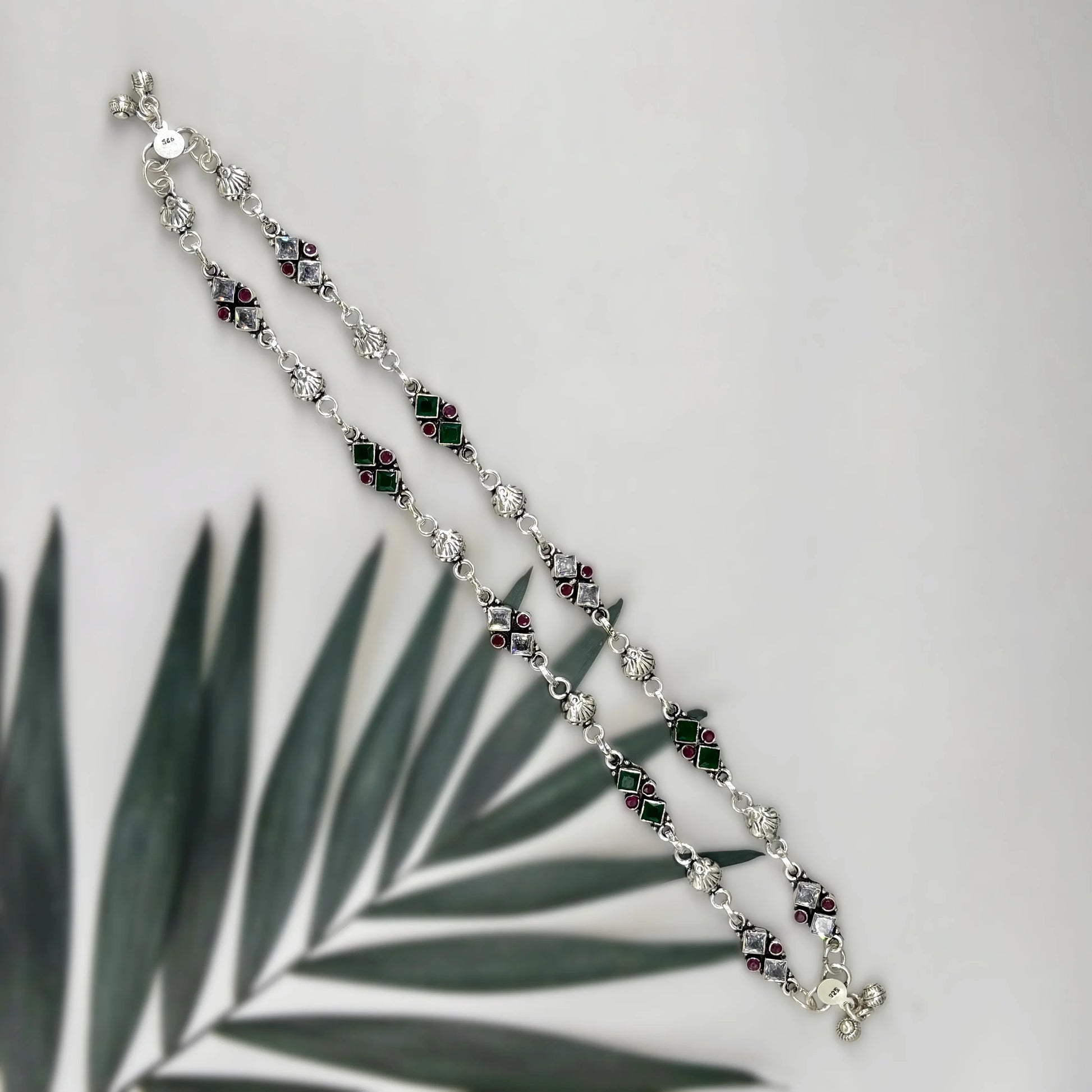 Silver anklet with green and red gemstones on white background with a palm leaf