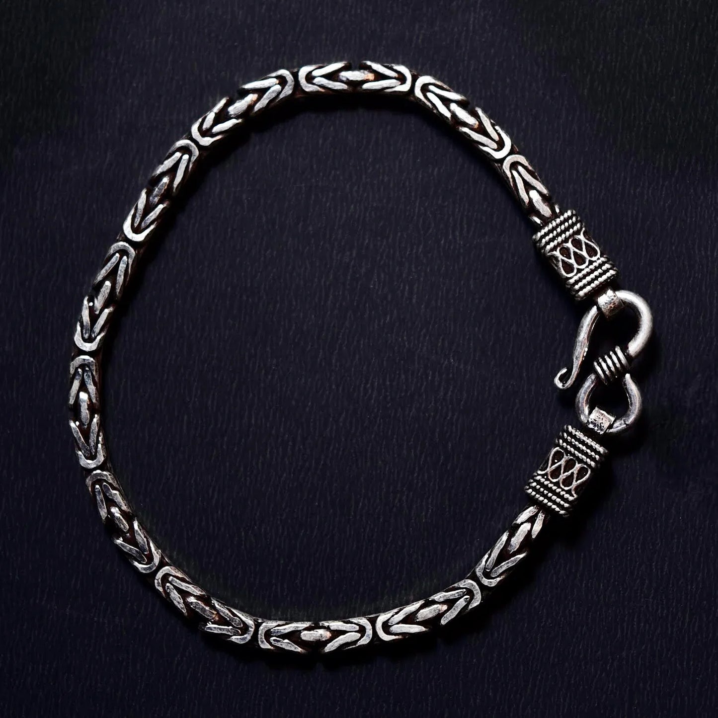 Silver chain bracelet with intricate woven pattern and decorative clasp on black background