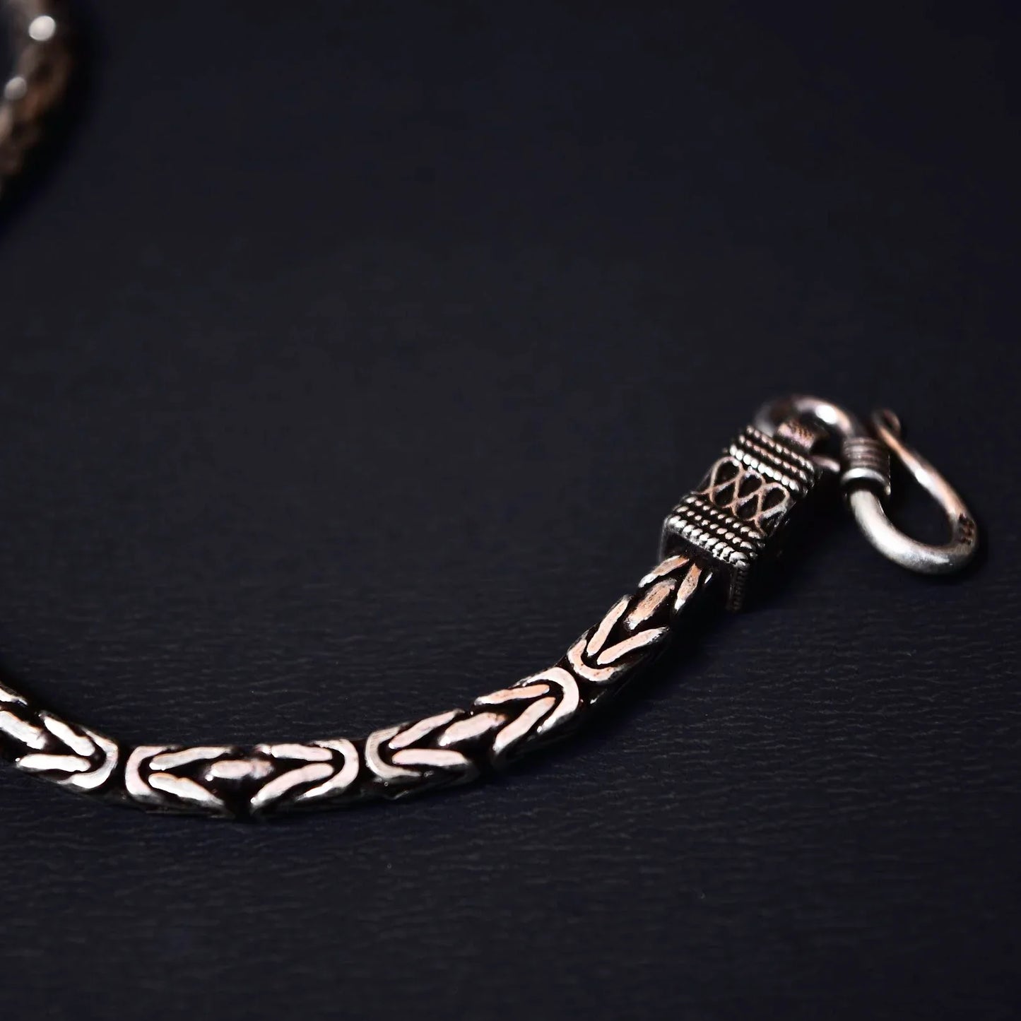 Close-up of a silver chain bracelet with intricate braided links and a hook clasp on a black background