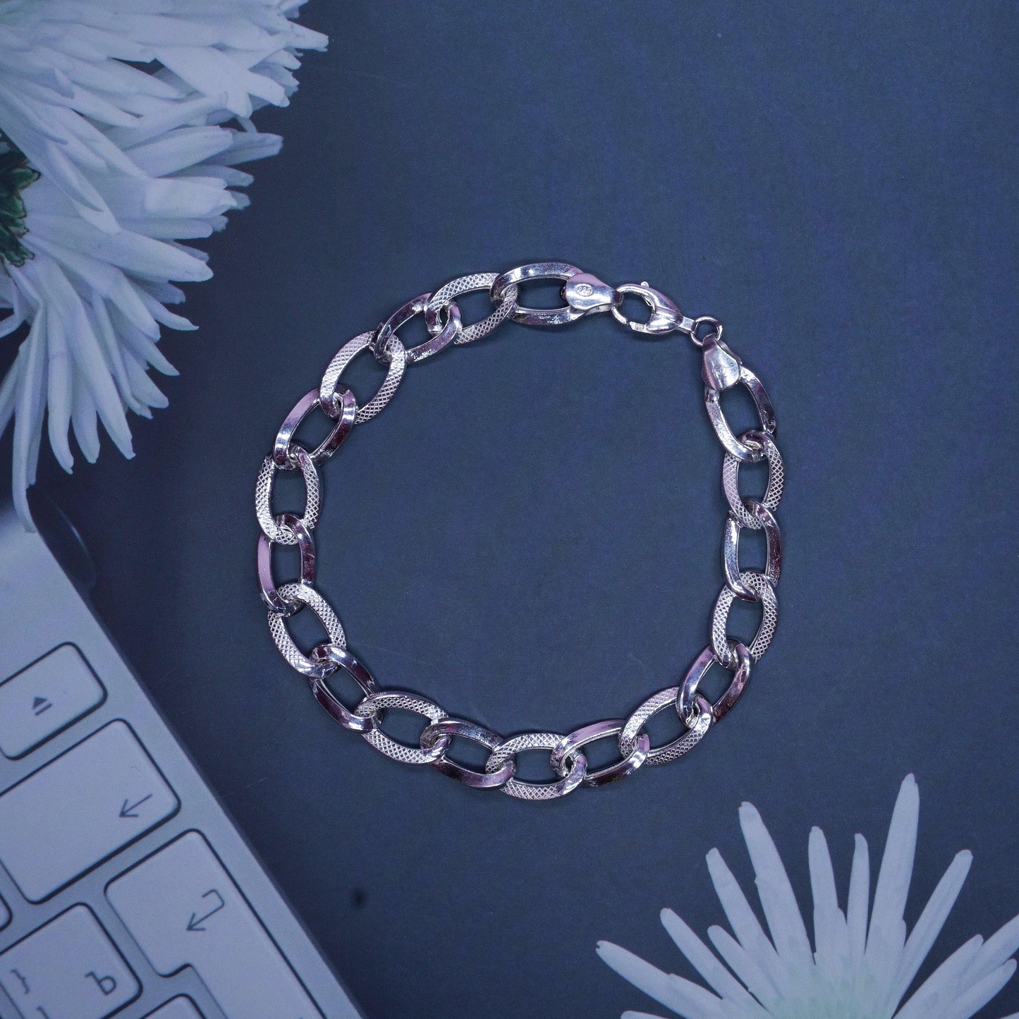 Silver chain link bracelet with textured and smooth oval links on dark background with white flowers and keyboard
