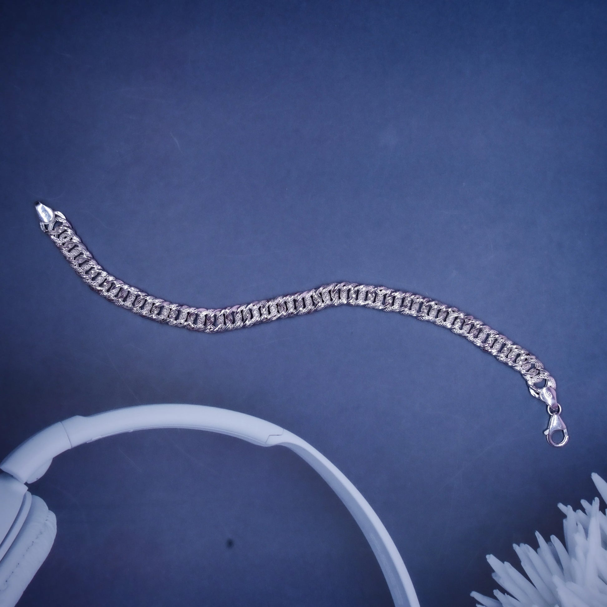 Silver chain bracelet with clasp on dark blue background near white headphones and flower