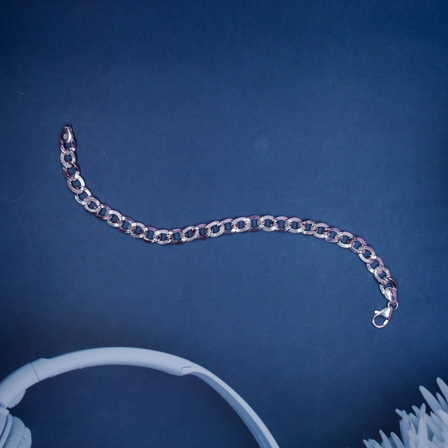 Silver chain bracelet with textured links on dark blue background beside white headphones and fluffy white fabric