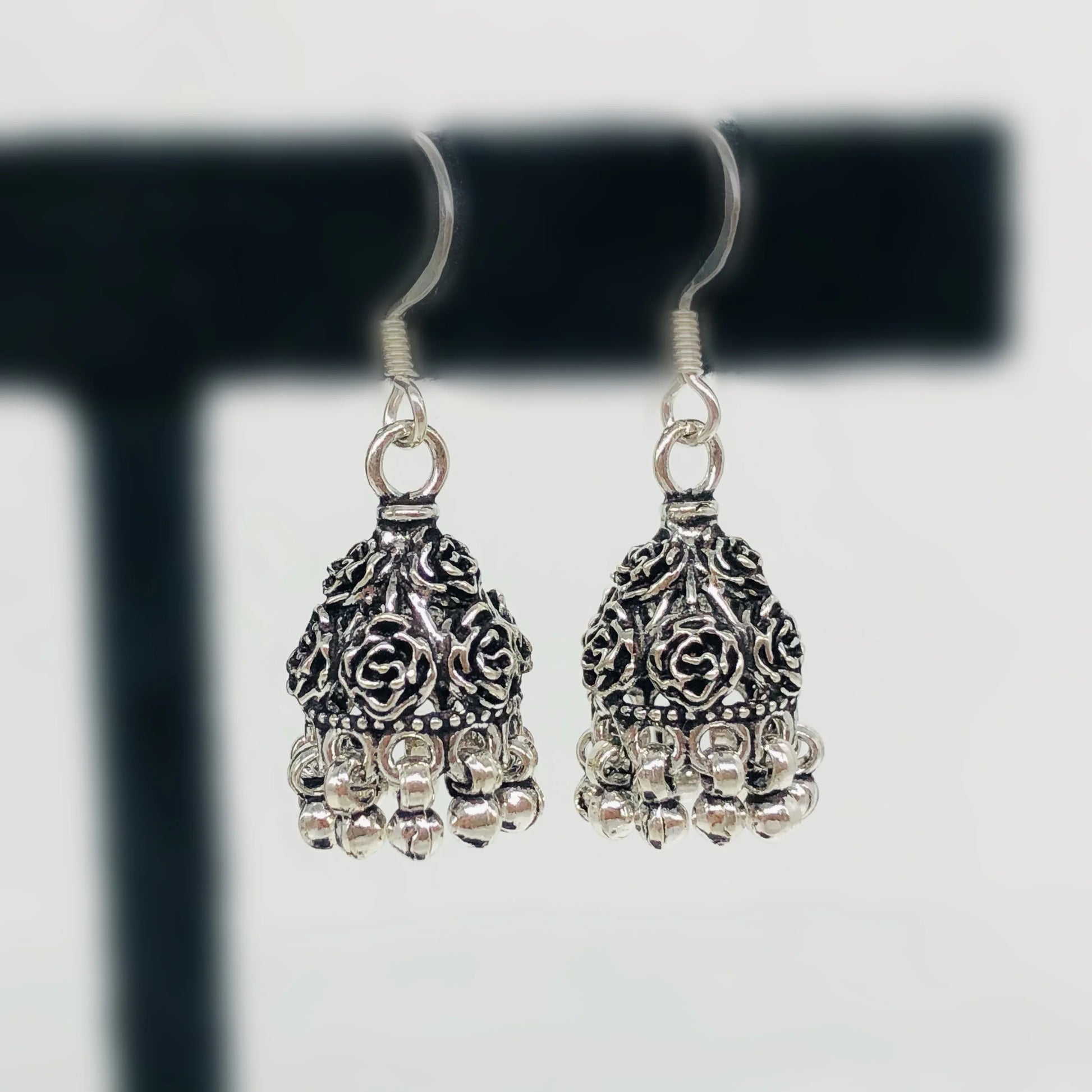 Antique silver jhumka earrings with floral design and dangling beads on black stand
